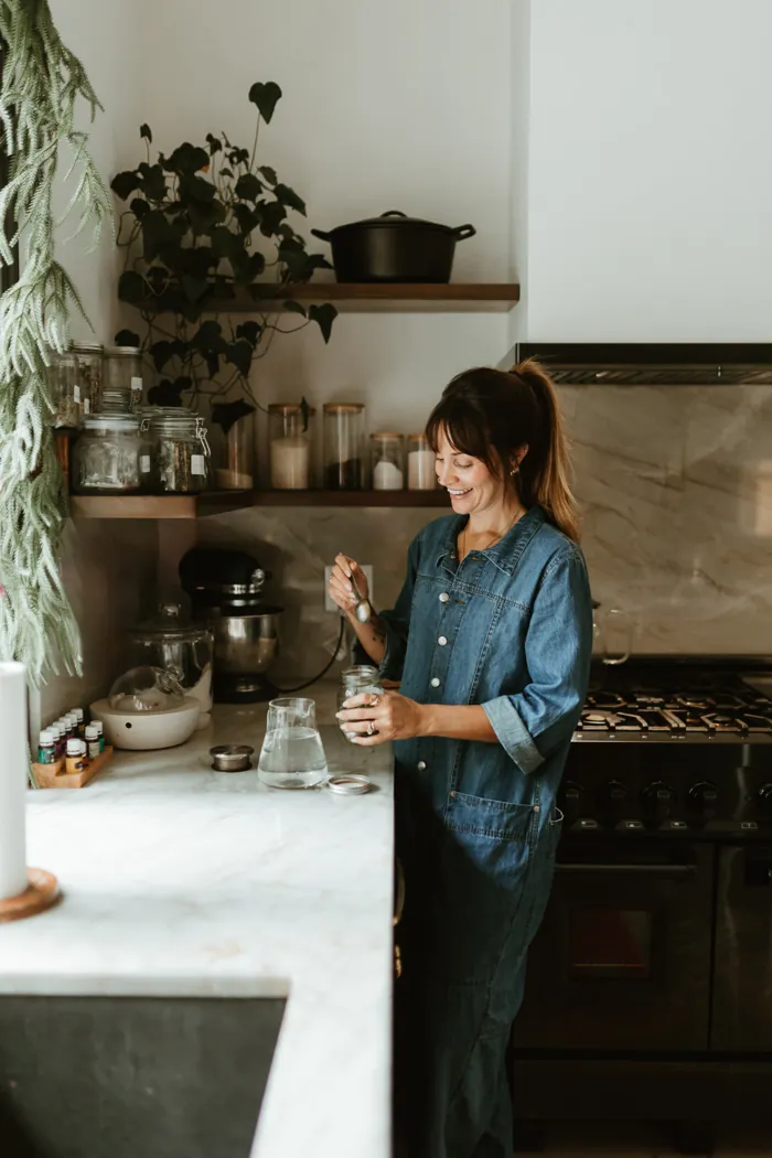 Jess Manuel in a denim jumpsuit stirring a jar at her kitchen counter